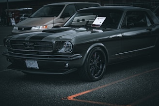A classic black Ford Mustang is parked in a lot. It has a sleek, shiny body with black rims and a visible license plate. A paper is placed on the dashboard. Another vehicle, a light brown minivan, is parked nearby in the background.