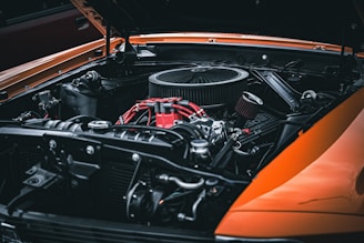 A close-up view of a car engine with a focus on the intricate details of the components. The engine is predominantly metallic with some parts in red, including wires and a section labeled '302'. The hood of the car is open, and the body is painted in a striking orange.