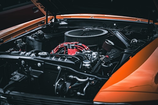 A close-up view of a car engine with a focus on the intricate details of the components. The engine is predominantly metallic with some parts in red, including wires and a section labeled '302'. The hood of the car is open, and the body is painted in a striking orange.