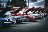 A line of classic muscle cars is parked outdoors with their hoods open, exposing the engines. The cars are an array of colors including blue, green, and red, and are set against a backdrop of trees and a cloudy sky, suggesting a car show or gathering.