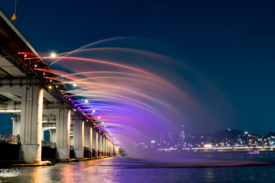 The image depicts a vibrant cityscape at night, with a large bridge spanning across a river and adorned with colorful lights that create an enchanting spectacle. The bridge is illuminated by purple, pink, and orange lights, casting a warm glow on the surrounding area. The water below mirrors the colors of the lights, creating a beautiful reflection. In the distance, the city skyline can be seen, with buildings of varying heights and designs, including a tall skyscraper that stands out against the night sky.