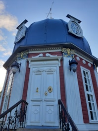 A small, ornate building featuring a blue domed roof with circular windows. The facade includes a grand white door adorned with golden decorations, framed by red and white paneling. Decorative black iron railings flank the entrance, and traditional lantern-style lamps are mounted on either side of the door.