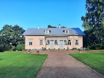 A quaint, two-story house with a pink facade and a blue-gray metal roof is surrounded by lush greenery. A symmetrical front of the house features several windows and a main door with decorative glass panels. There are potted plants lined neatly along the stone pathway leading up to the entrance. The foreground consists of a well-manicured lawn and a dirt pathway.