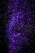 A vibrant cascade of purple particles, resembling a nebula or cosmic cloud, set against a deep black background. The particles are densely clustered and layered, creating a sense of depth and movement.