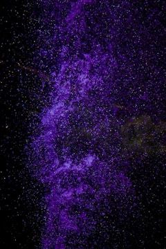 A vibrant cascade of purple particles, resembling a nebula or cosmic cloud, set against a deep black background. The particles are densely clustered and layered, creating a sense of depth and movement.