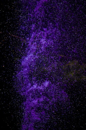A vibrant cascade of purple particles, resembling a nebula or cosmic cloud, set against a deep black background. The particles are densely clustered and layered, creating a sense of depth and movement.