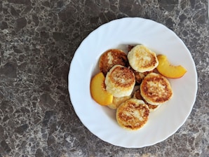 A white plate filled with small, round, golden brown pancakes arranged on a dark marbled surface. Alongside the pancakes, slices of fresh peach add a touch of color to the arrangement.