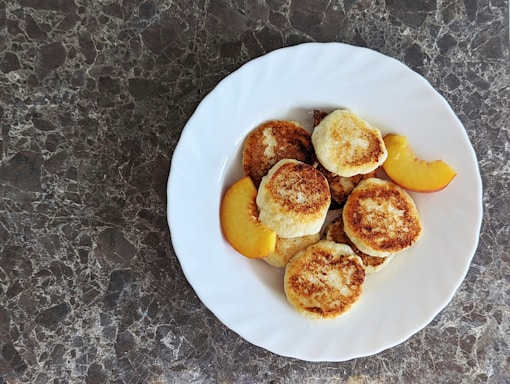 A white plate filled with small, round, golden brown pancakes arranged on a dark marbled surface. Alongside the pancakes, slices of fresh peach add a touch of color to the arrangement.