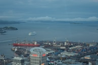 A bustling harbor scene with a large white cruise ship moving through the water. Numerous cranes and shipping containers are visible, indicating a busy port environment. Prominent buildings, including one with 'Vero' written on it, are visible in the foreground. The background shows a cloudy sky and distant islands.