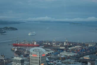 A bustling harbor scene with a large white cruise ship moving through the water. Numerous cranes and shipping containers are visible, indicating a busy port environment. Prominent buildings, including one with 'Vero' written on it, are visible in the foreground. The background shows a cloudy sky and distant islands.