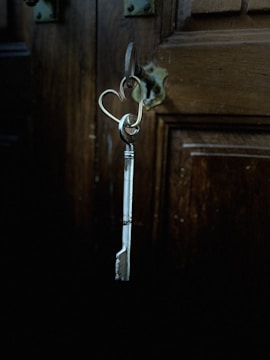 A metallic key hangs from a heart-shaped keyring inserted into a lock on a wooden door. The background is dimly lit, emphasizing the rustic texture and dark color of the wood.