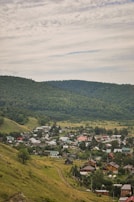 A small, picturesque village is nestled in a valley surrounded by lush, green hills under a partly cloudy sky. The village consists of numerous quaint houses with various colored roofs, predominantly red and green. A dirt path winds through the grassy landscape leading towards the forested area in the distance. The setting conveys a sense of peaceful, rural living among nature.