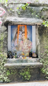 A small shrine is set into a rock wall, displaying a colorful painting of Hindu deities with offerings such as flowers and small statues placed in front. Green foliage and vines partially cover the surrounding stone surface.