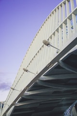 An architectural structure with a series of vertical white metallic beams and overhead lights, showcasing a modern bridge design under a clear blue sky.