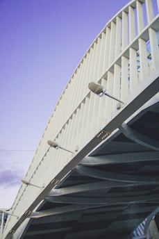 An architectural structure with a series of vertical white metallic beams and overhead lights, showcasing a modern bridge design under a clear blue sky.