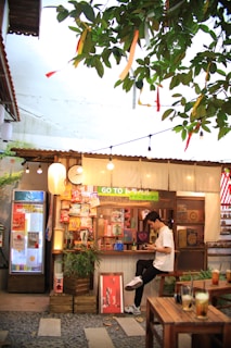 A cozy outdoor cafe features a small wooden booth adorned with various snacks and beverages. A young man sits on a stool, absorbed in his smartphone, with a relaxed demeanor. Above, decorative ribbons and warm string lights hang from lush green tree branches, creating a vibrant and inviting atmosphere.