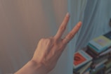 A hand is making a peace sign gesture with two fingers extended. The background is softly blurred, showing a stack of books with various colored covers. The hand wears a couple of rings on the middle finger.