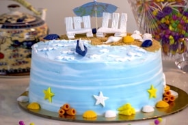 A beautifully decorated cake with a beach theme, featuring blue icing resembling ocean waves, decorated with miniature seashells and starfish. The top of the cake is designed to look like sand with small white chairs and a beach umbrella. Surrounding the cake are various colorful decorations, including small shells and stars in yellow and orange hues. A teapot with a detailed design and a colorful glass container of candies are visible in the background.