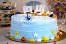 A beautifully decorated cake with a beach theme, featuring blue icing resembling ocean waves, decorated with miniature seashells and starfish. The top of the cake is designed to look like sand with small white chairs and a beach umbrella. Surrounding the cake are various colorful decorations, including small shells and stars in yellow and orange hues. A teapot with a detailed design and a colorful glass container of candies are visible in the background.