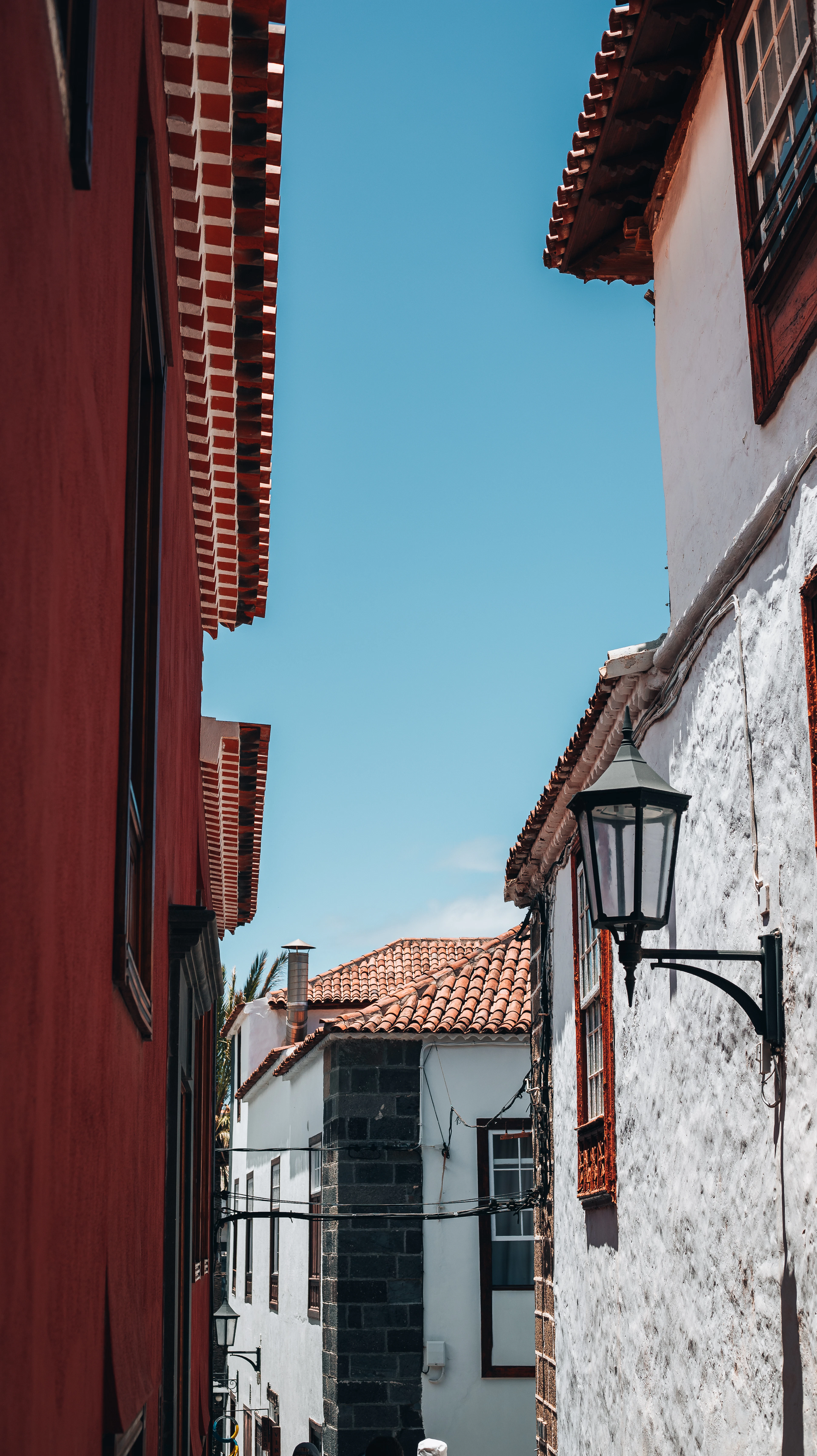 A narrow lane in Garachico, Tenerife, hemmed by a deep red wall and a whitewashed house with dark volcanic stone corners, a wrought-iron lantern and terracotta-tiled eaves cutting into a clear blue sky.