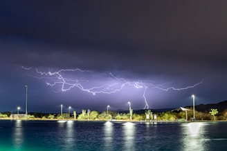 A dramatic lightning storm is happening over a body of water at night. The sky is illuminated with multiple lightning bolts, and the water reflects the light from the street lamps and lightning. Trees and bushes are visible along the shore, and street lamps are evenly spaced, providing light to the area. Some structures, including a small building and signage, can be seen in the background.