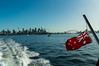 A city skyline is visible across the water under a clear blue sky, with a prominent bridge in the distance. The image captures a sense of motion with water trails in the foreground, suggesting the picture was taken from a moving boat. An Australian flag flutters in the breeze, adding a patriotic element to the scene.