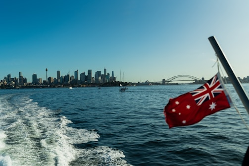 A city skyline is visible across the water under a clear blue sky, with a prominent bridge in the distance. The image captures a sense of motion with water trails in the foreground, suggesting the picture was taken from a moving boat. An Australian flag flutters in the breeze, adding a patriotic element to the scene.