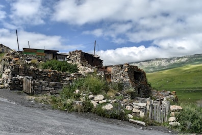 A rustic stone building with an aged, weathered appearance is set against a backdrop of rolling green hills and a cloudy blue sky. The structure includes a wooden gate and is surrounded by a variety of greenery, including shrubs and grass. A sign, partially visible, suggests the building may serve as some type of lodge or hostel.