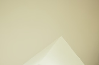 A minimalistic image featuring the corner of a triangular paper against a smooth, light beige background. The sharp angles and soft color tones create a sense of simplicity.