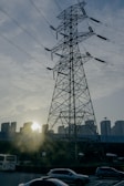 A large power transmission tower stands prominently against a backdrop of urban buildings. The sun is setting behind the buildings, casting a warm glow and creating silhouettes of the structures. In the foreground, several cars are on a road, indicating an urban setting.