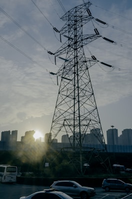 A large power transmission tower stands prominently against a backdrop of urban buildings. The sun is setting behind the buildings, casting a warm glow and creating silhouettes of the structures. In the foreground, several cars are on a road, indicating an urban setting.