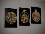 Three black canvases with intricate Arabic calligraphy in gold, each featuring unique ornate designs. The calligraphy is presented in different symmetrical shapes, with the middle canvas having a pointed arch-like form.
