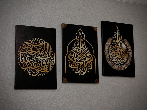 Three black canvases with intricate Arabic calligraphy in gold, each featuring unique ornate designs. The calligraphy is presented in different symmetrical shapes, with the middle canvas having a pointed arch-like form.