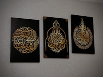 Three black canvases with intricate Arabic calligraphy in gold, each featuring unique ornate designs. The calligraphy is presented in different symmetrical shapes, with the middle canvas having a pointed arch-like form.