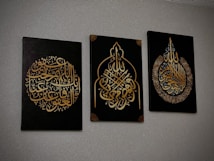 Three black canvases with intricate Arabic calligraphy in gold, each featuring unique ornate designs. The calligraphy is presented in different symmetrical shapes, with the middle canvas having a pointed arch-like form.