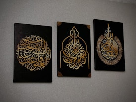 Three black canvases with intricate Arabic calligraphy in gold, each featuring unique ornate designs. The calligraphy is presented in different symmetrical shapes, with the middle canvas having a pointed arch-like form.