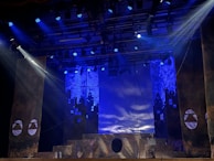 A theatrical stage setup with dramatic lighting, featuring large pillars and a geometric design backdrop. Blue and purple lights create a moody atmosphere, while ropes and stage structures add depth to the scene.