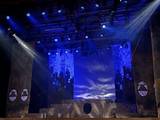 A theatrical stage setup with dramatic lighting, featuring large pillars and a geometric design backdrop. Blue and purple lights create a moody atmosphere, while ropes and stage structures add depth to the scene.