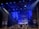 A theatrical stage setup with dramatic lighting, featuring large pillars and a geometric design backdrop. Blue and purple lights create a moody atmosphere, while ropes and stage structures add depth to the scene.