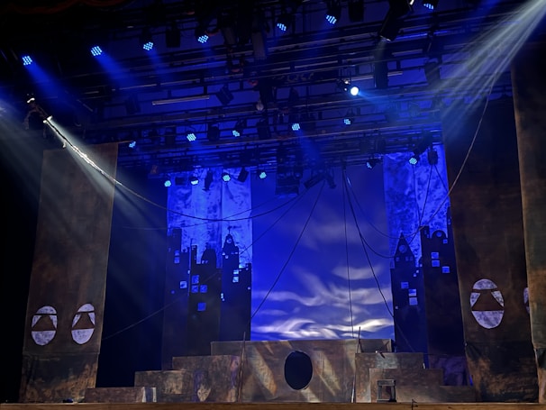 A theatrical stage setup with dramatic lighting, featuring large pillars and a geometric design backdrop. Blue and purple lights create a moody atmosphere, while ropes and stage structures add depth to the scene.