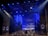 A theatrical stage setup with dramatic lighting, featuring large pillars and a geometric design backdrop. Blue and purple lights create a moody atmosphere, while ropes and stage structures add depth to the scene.