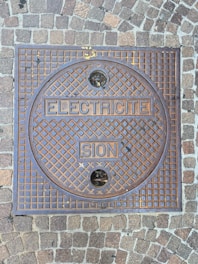 A square metal manhole cover set within a sidewalk made of cobblestone pavers. The manhole cover has a grid pattern with the words 'ELECTRICITE' and 'SION' embossed on it. Two small circular holes are present in the upper and lower sections of the cover.