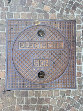 A square metal manhole cover set within a sidewalk made of cobblestone pavers. The manhole cover has a grid pattern with the words 'ELECTRICITE' and 'SION' embossed on it. Two small circular holes are present in the upper and lower sections of the cover.