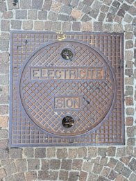 A square metal manhole cover set within a sidewalk made of cobblestone pavers. The manhole cover has a grid pattern with the words 'ELECTRICITE' and 'SION' embossed on it. Two small circular holes are present in the upper and lower sections of the cover.