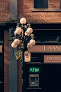 A vintage street lamp with spherical glass bulbs is mounted on a metal post. The post also supports a decorative hanging basket with flowers. Below the lamp, there is a neon green ATM sign and an actual ATM machine embedded in a brick wall. To the left of the ATM, there is a no-parking sign. The background consists of the brick façade of a building, with a small window above the ATM, and partial signage for a business establishment is visible.
