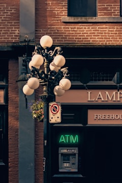 A vintage street lamp with spherical glass bulbs is mounted on a metal post. The post also supports a decorative hanging basket with flowers. Below the lamp, there is a neon green ATM sign and an actual ATM machine embedded in a brick wall. To the left of the ATM, there is a no-parking sign. The background consists of the brick façade of a building, with a small window above the ATM, and partial signage for a business establishment is visible.