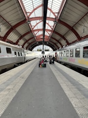 A train station platform flanked by two stationary trains under an arched, red and white metal roof. A group of people with luggage sits and stands in the center, waiting. The platform is lined with tactile paving, and the station is well-lit by natural light from large windows.