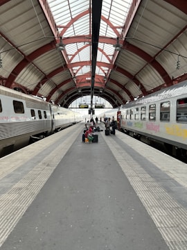 A train station platform flanked by two stationary trains under an arched, red and white metal roof. A group of people with luggage sits and stands in the center, waiting. The platform is lined with tactile paving, and the station is well-lit by natural light from large windows.