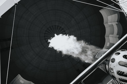 The image captures the interior of a hot air balloon from the perspective inside the balloon's envelope. A burner is releasing a blast of flame upwards, inflating the balloon. The fabric of the balloon envelops the space, and the concentric lines create a dramatic focal point.
