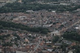 Aerial view of a city with dense historical architecture, featuring church steeples and a mix of modern buildings surrounded by lush green areas. Red and brown rooftops dominate the landscape, and wide streets intersect through the cityscape.